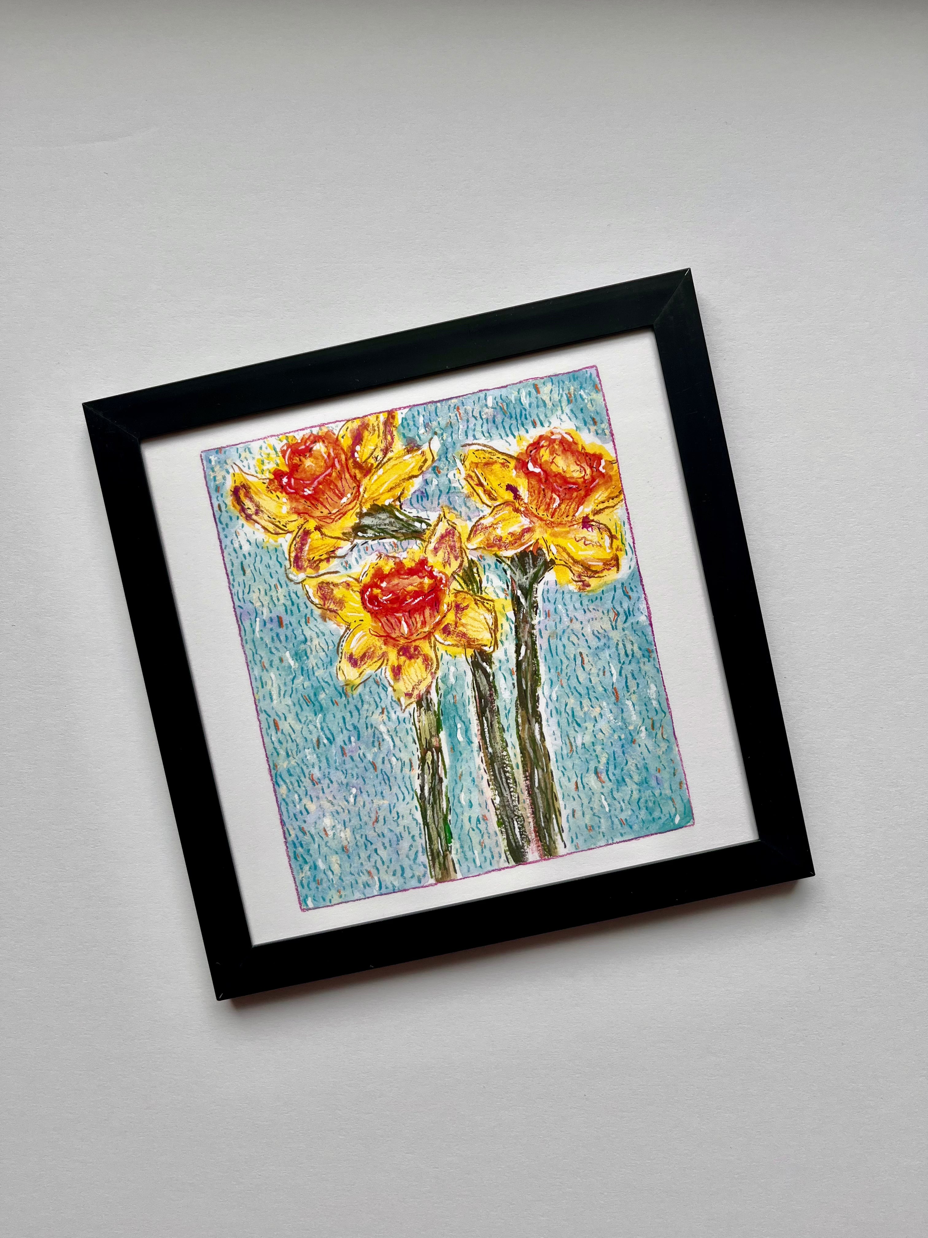 A framed artwork depicting three yellow and orange daffodils with green stems on a textured blue background, presented in a black frame with a white mat against a light gray background.